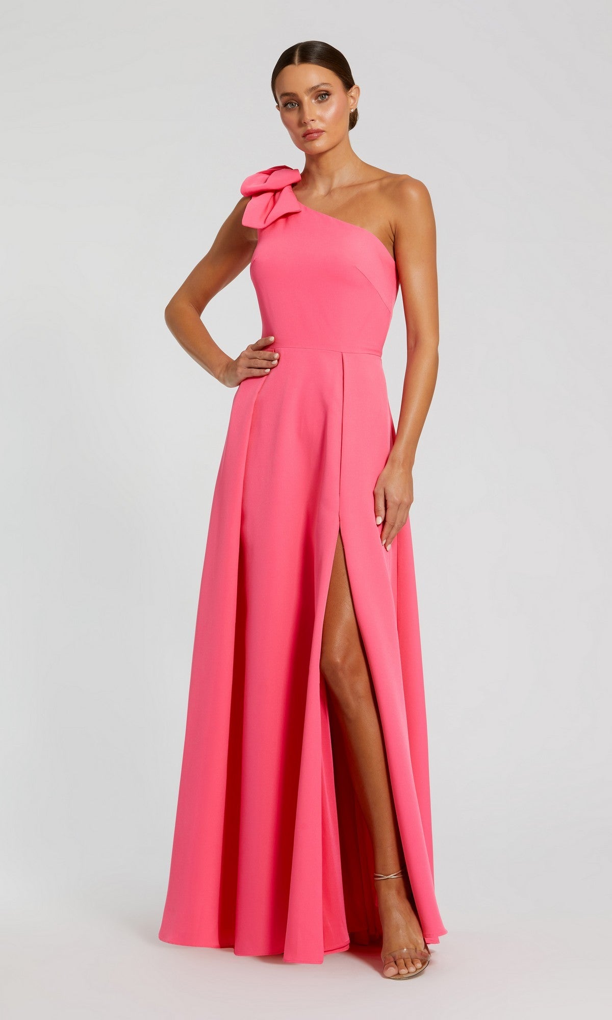 A woman wearing the Mac Duggal 26989 Long Formal Dress in candy pink—a floor-length, one-shoulder gown featuring a bow detail and high thigh slit—stands against a plain light background.